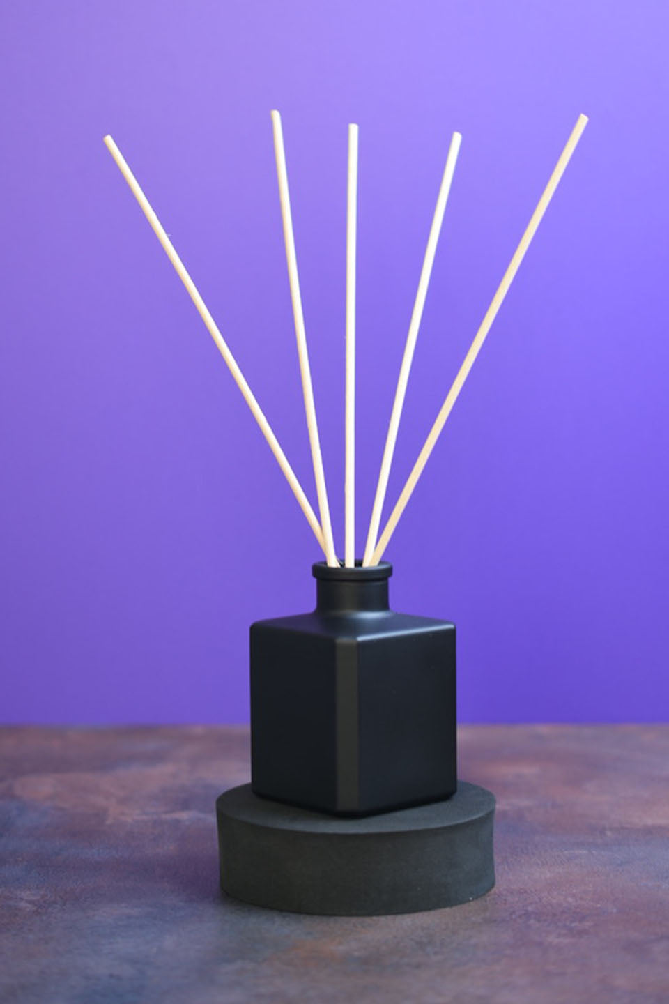 Matte black square reed diffuser bottle with 5 reeds sticking out of it. The bottle sits upon a black riser in front of a purple background.