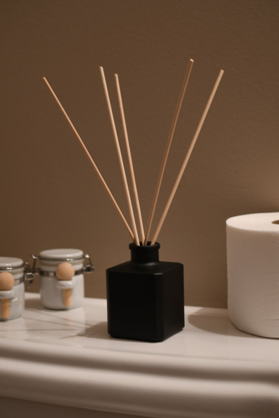 Matte Black square diffuser bottle with 5 reeds sticking out of it in a natural home bathroom setting.