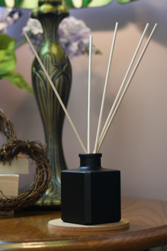 Matte black, square diffuser bottle with 5 reeds sticking out of it in upon a wood table with a stainglass lamp in the background.