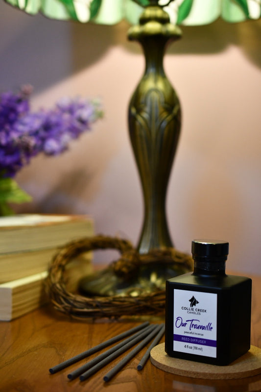 Collie Creek Candles reed diffuser bottle on a side table with gray reeds alongside the bottle, a group of lilac flowers appear in the background.