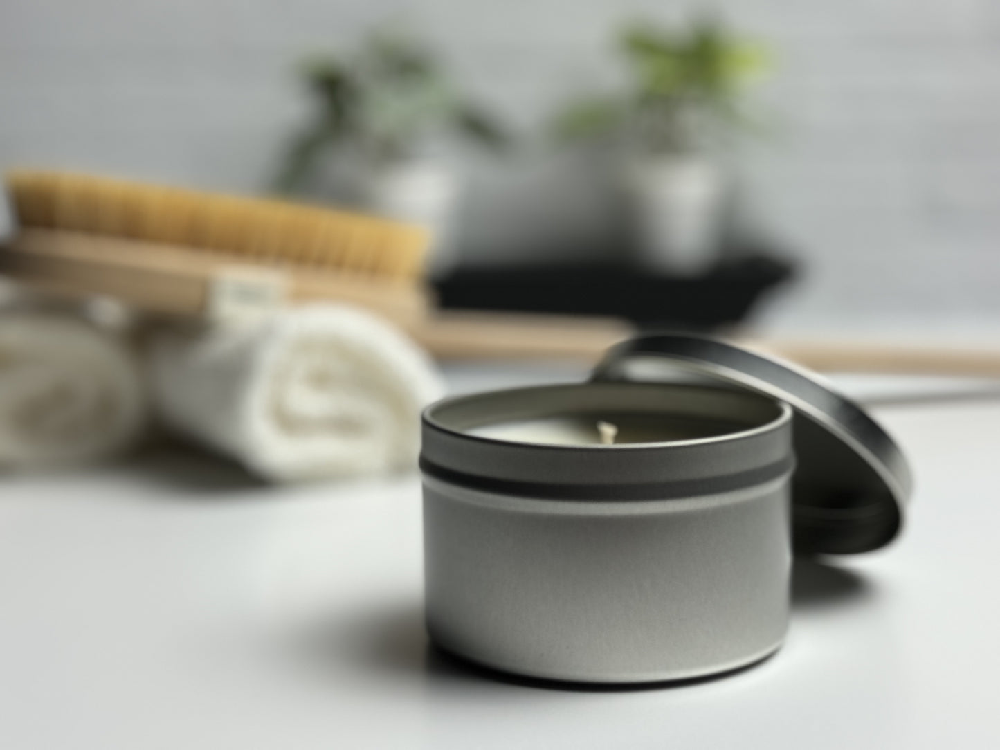 Tin candle in spa like setting, rolled towels, a body brush and green potted plants in the background.