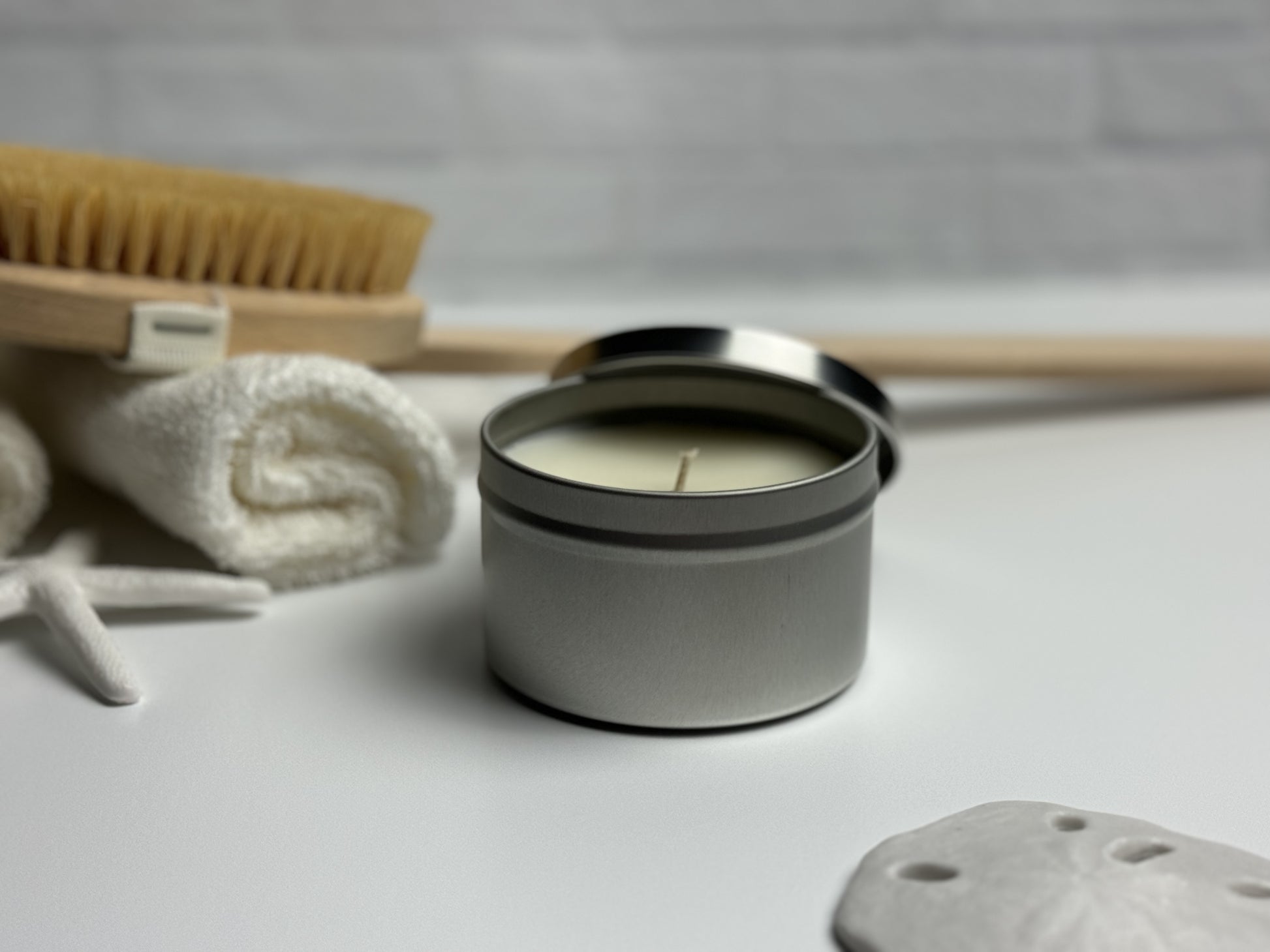Tin candle in spa like setting, sea shell in foreground and starfish near rolled towels and a body brush in the background.
