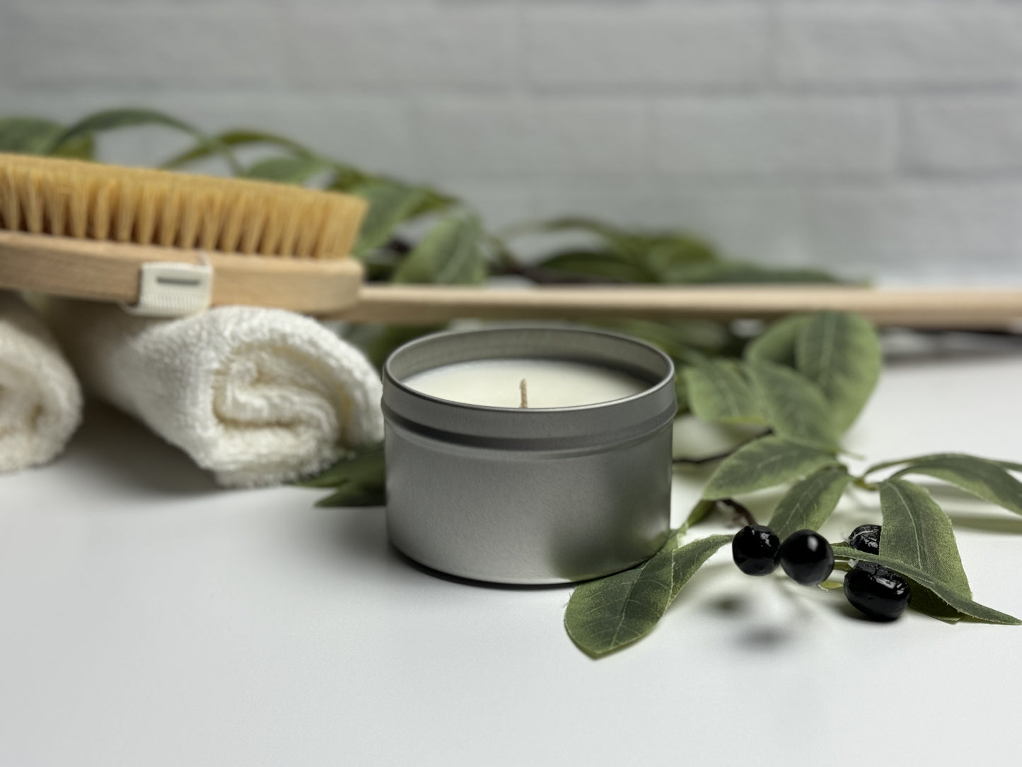 Tin candle in spa like setting. Olive branch adorns the foreground with rolled towels and a body brush in the background.
