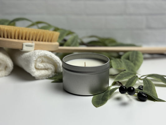 Tin candle in spa like setting. Olive branch adorns the foreground with rolled towels and a body brush in the background.