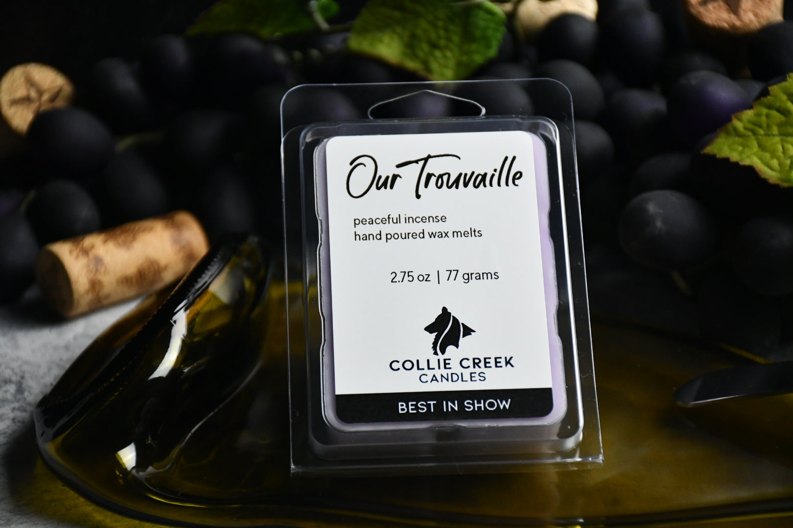 Our Trouvaille wax melt sets upon a flattened, green glass wine bottle amidst grapes and vines with scattered wine corks.