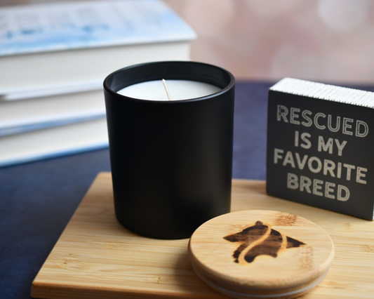 A matte black, single wicked candle in a home setting. Books are blurred in the background and a "Rescued is my favorite breed" sign and candle lid appear in the foreground.