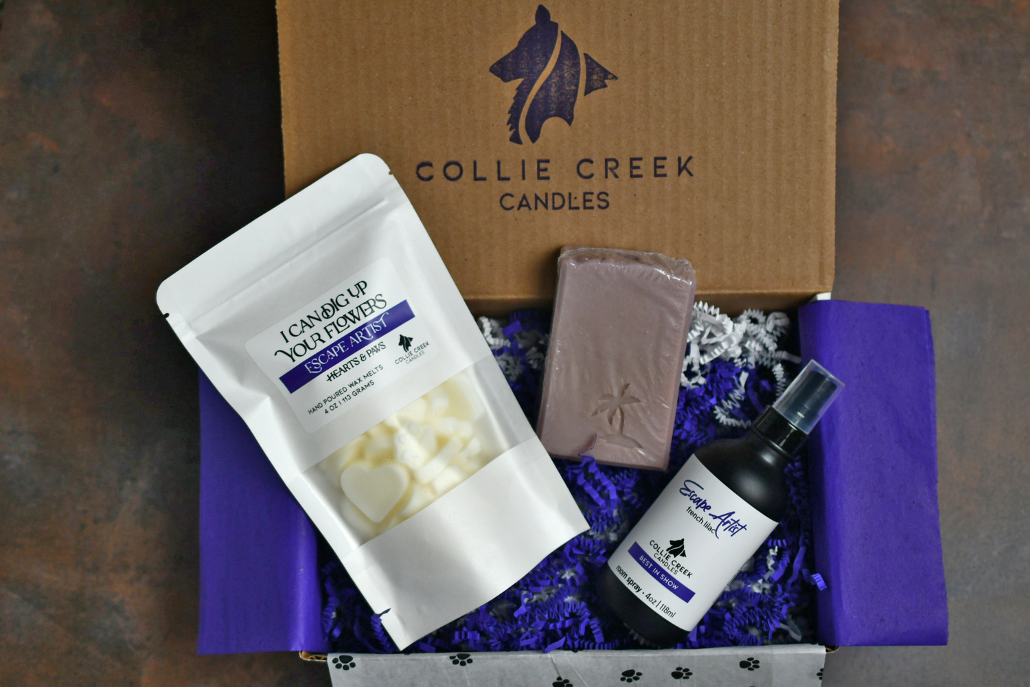 Box set with pouch of wax melts, bar of lilac soap and glass bottle of room spray on a bed of purple and white crinkle paper.