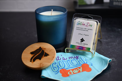 Indigo glass vessel, single wick candle and 6-cell clam shell of Gotcha Day scented wax melt on a black surface with a dog bandana that reads "it's my gotcha day" in the foreground.