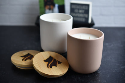 Two ceramic candles with wooden lids, arranged on a black marbled surface. One is blush colored and the other is white. A wood burned logo design on the lids sits in the foreground.