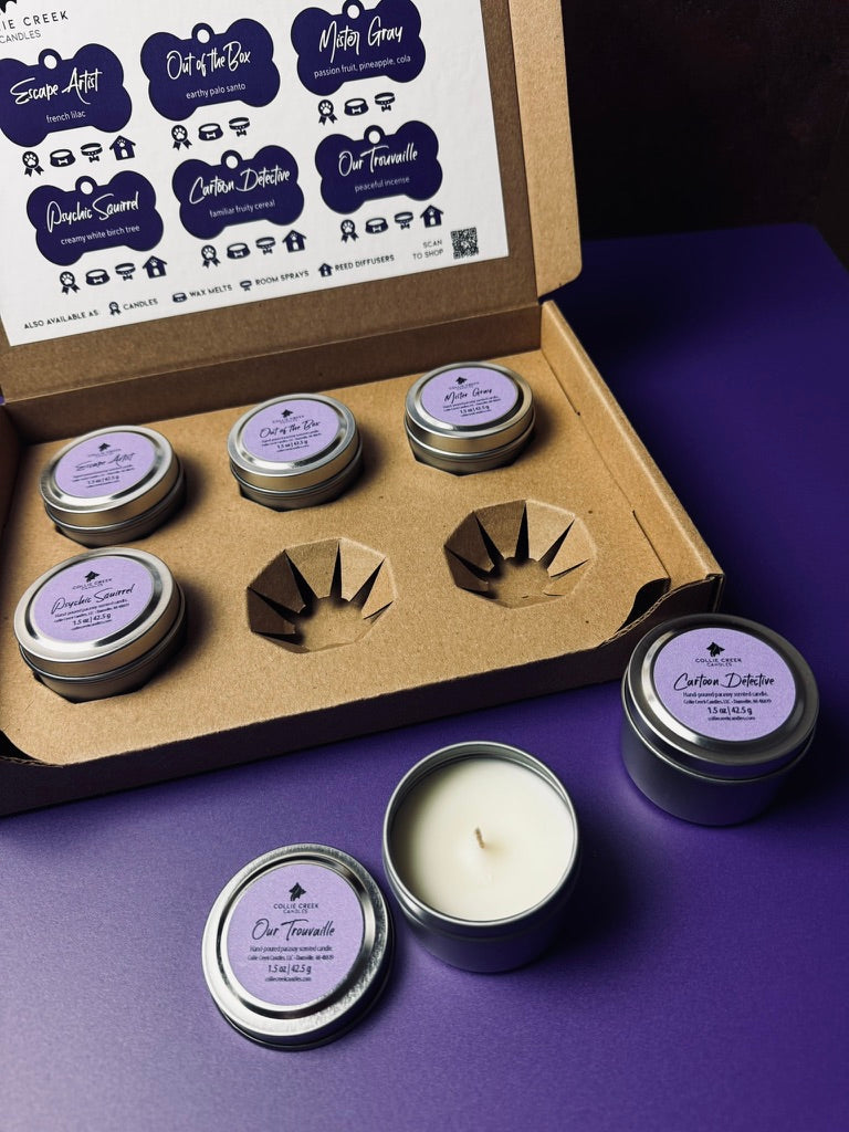 Set of candles with purple labels in a cardboard box on a purple surface one candle has the lid removed to show a single wick in natural-colored wax.