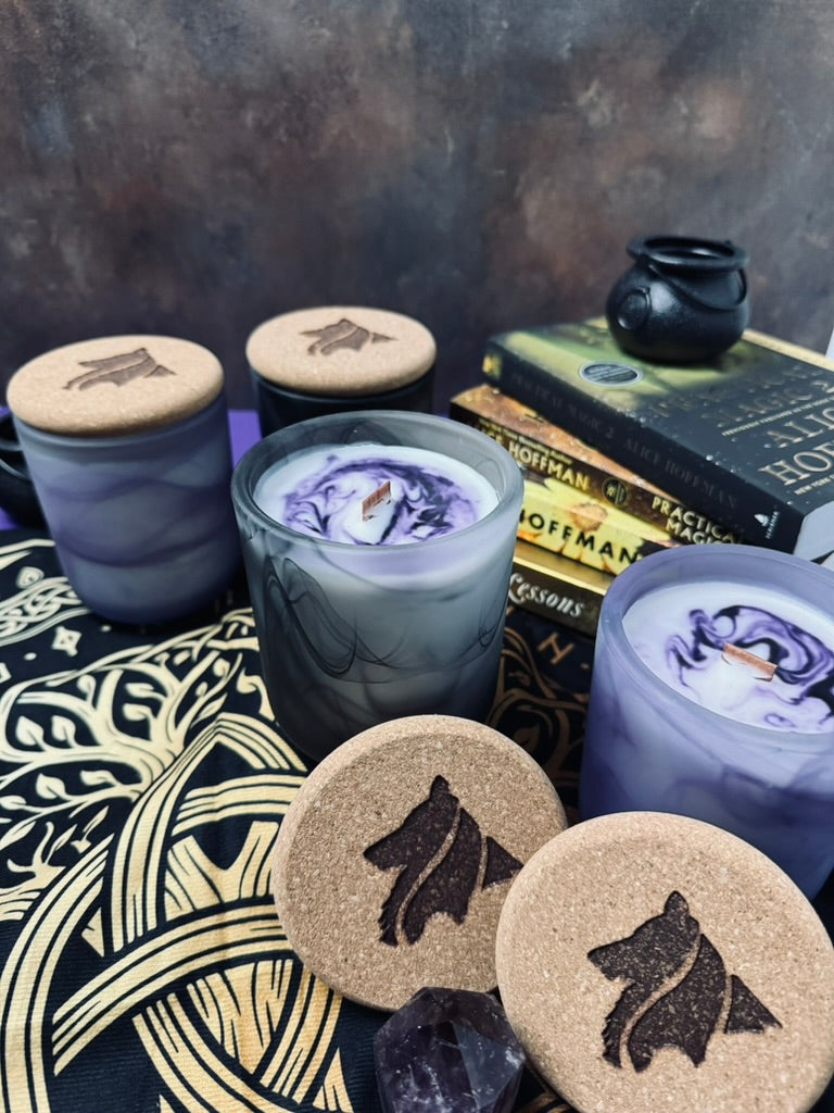Collie Creek Candles logo burned onto cork tops in the foreground with 2 purple swirl candles and 2 black swirl candles sit in front of a stack of books by Alice Hoffman that inspired the collection.