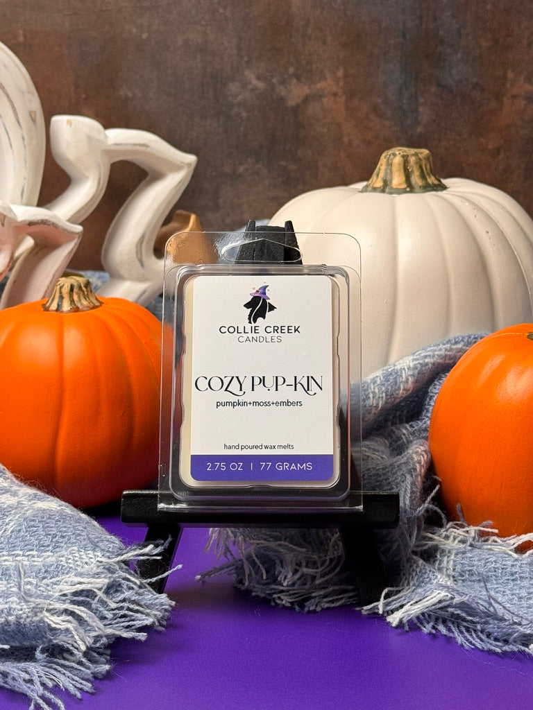 Collie Creek Cozy Pup-kin wax melt surrounded by decorative pumpkins and a soft blue woven scarf on a purple surface.