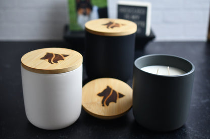 Three ceramic candles with wooden lids,  arranged on a dark marbled surface. One is a black color, another is gray and the other is white, with a wood burned logo design on the top.