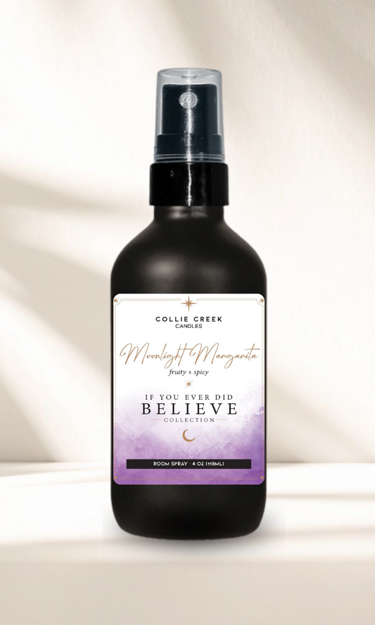 A black spray bottle of Collie Creek Candle 'Moonlight Margarita' room spray, featuring a white and purple label, set against a warm beige background with soft shadows.