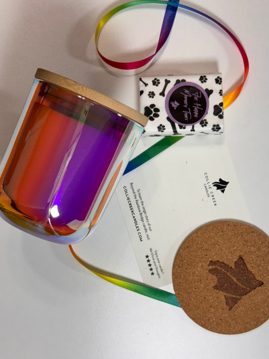 Rainbow colored jar candle with wooden lid, rainbow ribbon, and coaster on a white surface
