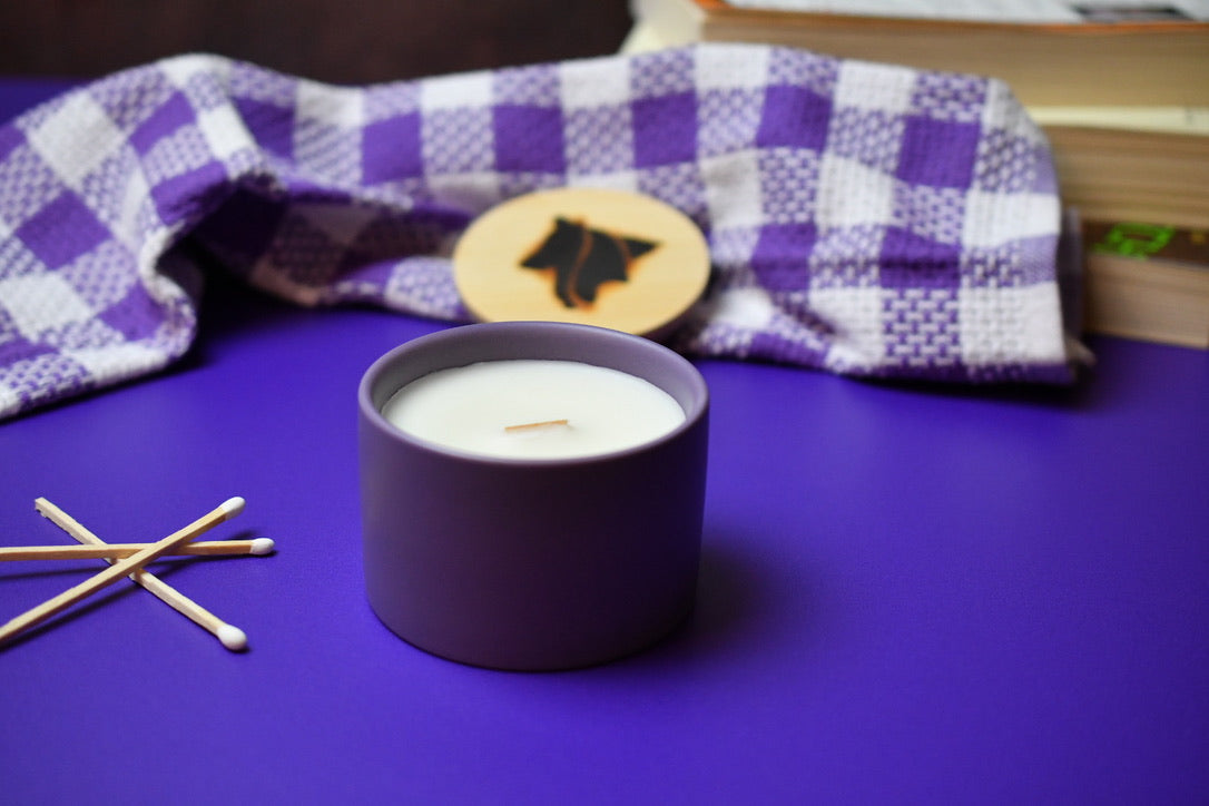 Candle in a purple container with matches on a purple surface, checkered cloth in the background