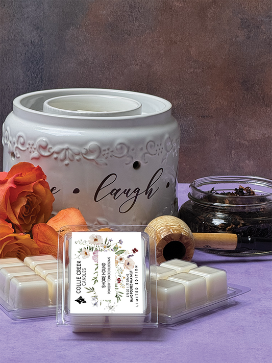 Natural colored wax melts in 6 cube clam shell. Floral designed label, 2.75 oz, 77 grams, limited edition. Set in display with peach colored roses, a corn cob pipe and a glass container of loose pipe tobacco on light purple background.