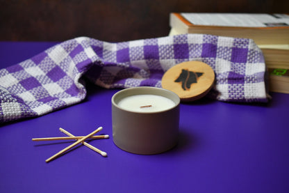 Candle in a stone container with wooden matches on a purple surface, next to a checkered cloth and books.