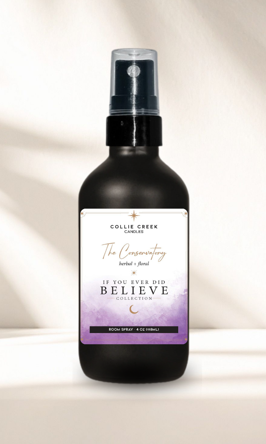 A matte black bottle of Collie Creek Candles' The Conservatory room spray with a clear spray top and a white label featuring purple and gold text, set against a warm off-white background with soft shadows.