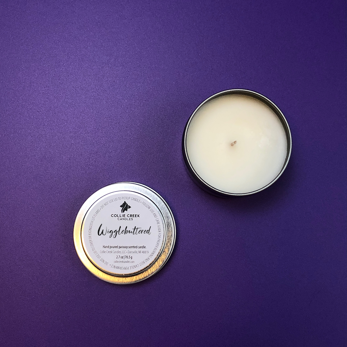 A silver tinned candle upon a purple background. The silver top is off the vessel and reads Wigglebuttered as the scent name.