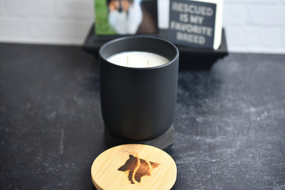 A black ceramic candle with wooden lid, arranged on a black marbled surface. A wood burned logo design on the lid sits in the foreground.