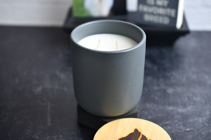 A gray ceramic candle with wooden lid, arranged on a black marbled surface. A wood burned logo design on the lid sits in the foreground.