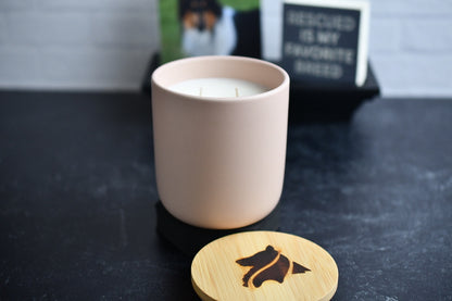 A blush colored ceramic candle with wooden lid, arranged on a black marbled surface. A wood burned logo design on the lid sits in the foreground.