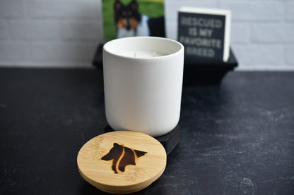 A white ceramic candle with wooden lid, arranged on a black marbled surface. A wood burned logo design on the lid sits in the foreground.