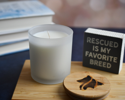 A white frosted, single wicked candle in a home setting. Books are blurred in the background and a "Rescued is my favorite breed" sign and candle lid appear in the foreground.